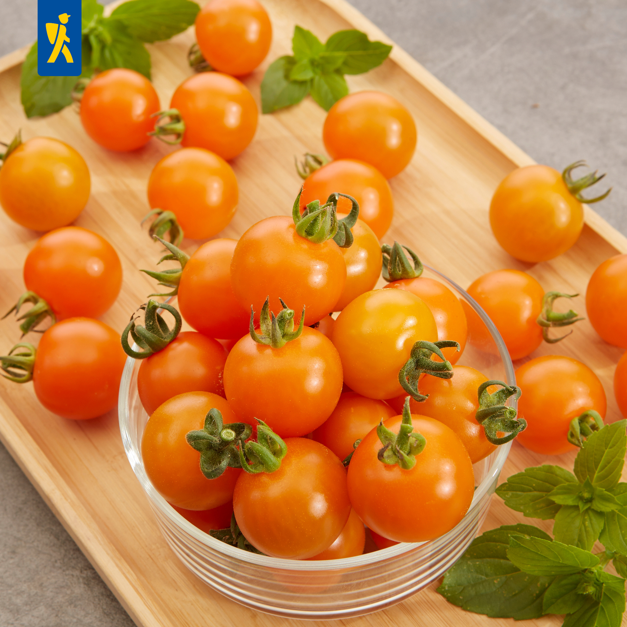 Orange Kirschtomaten in Glasschale und auf Holztablett, daneben frische grüne Blätter.