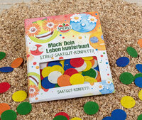 Bunte Streugut-Konfetti in einer Verpackung mit lachendem Gesicht und Blumen, auf Holzspänen verteilt.